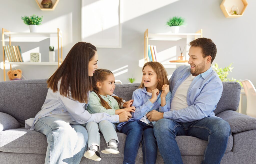 Happy smiling young family of four with two kids boy and girl sitting on sofa at home having fun and tickling resting on weekend. 
